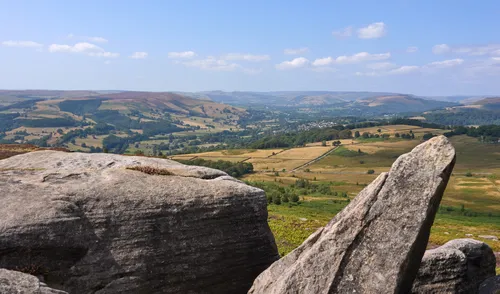 This landscape photograph captures a sweeping view of Hope Valley from atop Millstone Edge, situated within the Longshaw Estate in the Peak District of England, United Kingdom. The image was taken in the early afternoon during the summer season, as indicated by the bright sunlight and the lush greenery that dominates the valley and distant hills. The foreground prominently features rugged rocks characteristic of Millstone Edge, adding texture and depth to the scene. Rolling hills, scattered trees, and patchwork fields stretch out across the Hope Valley, offering a quintessential glimpse of the English countryside. The photograph showcases the natural beauty of the Peak District with its expansive views and summer vitality.